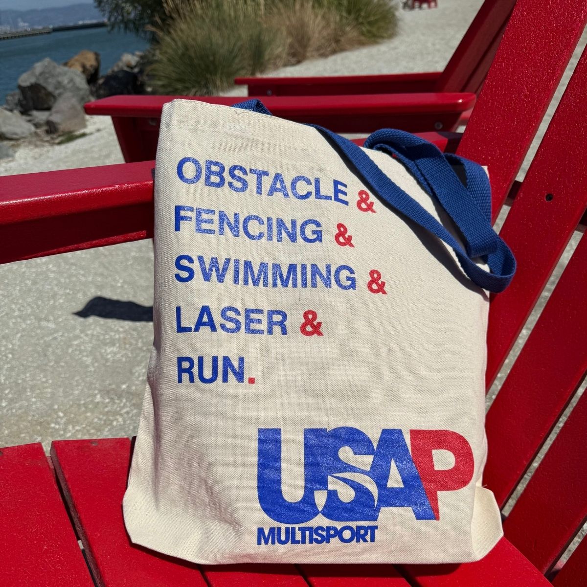USA Pentathlon Multisport tote bag listing the five events, resting on a red chair at the beach.